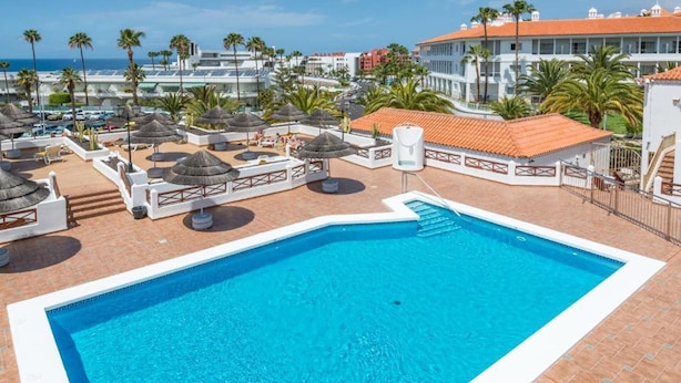 Piscina con borde blanco, rodeada de sillones de descanso y sombrillas, con vista al mar y edificios al fondo.