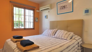 Un dormitorio con una cama grande, una ventana con rejas, un equipo de aire acondicionado y un cuadro enmarcado en la pared.