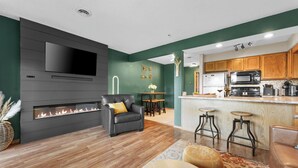 A modern living room with a fireplace, a TV mounted on the wall, and a kitchen area with wooden cabinets and a microwave. - Cozy Romance Vibes! 1 mile from Silver Dollar City-2 Pools-Free Boat Parking  (Reeds Spring, Mo)