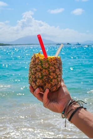 A hand holding a pineapple with a straw, set against a beach and ocean backdrop.