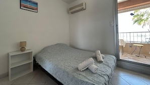 A single bed with a patterned bedspread, a small shelf with a lamp, and a picture on the wall. - Charming studio with sea view, Gruissan (Gruissan)