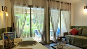A room with a sofa, a table with a book, and a view of an outdoor area with chairs and greenery. - Apartment with pool near the sea (Sanary-sur-Mer)