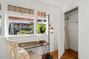 Um quarto com uma escrivaninha de madeira, uma cadeira, um abajur, uma planta em vaso e vista para um edifício pela janela.