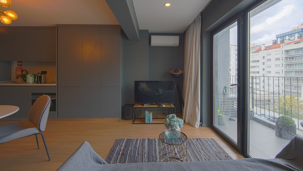 Ein modernes Wohnzimmer mit einer Couch, einem Couchtisch, einem Fernseher und einem Balkon mit Blick auf Gebäude.