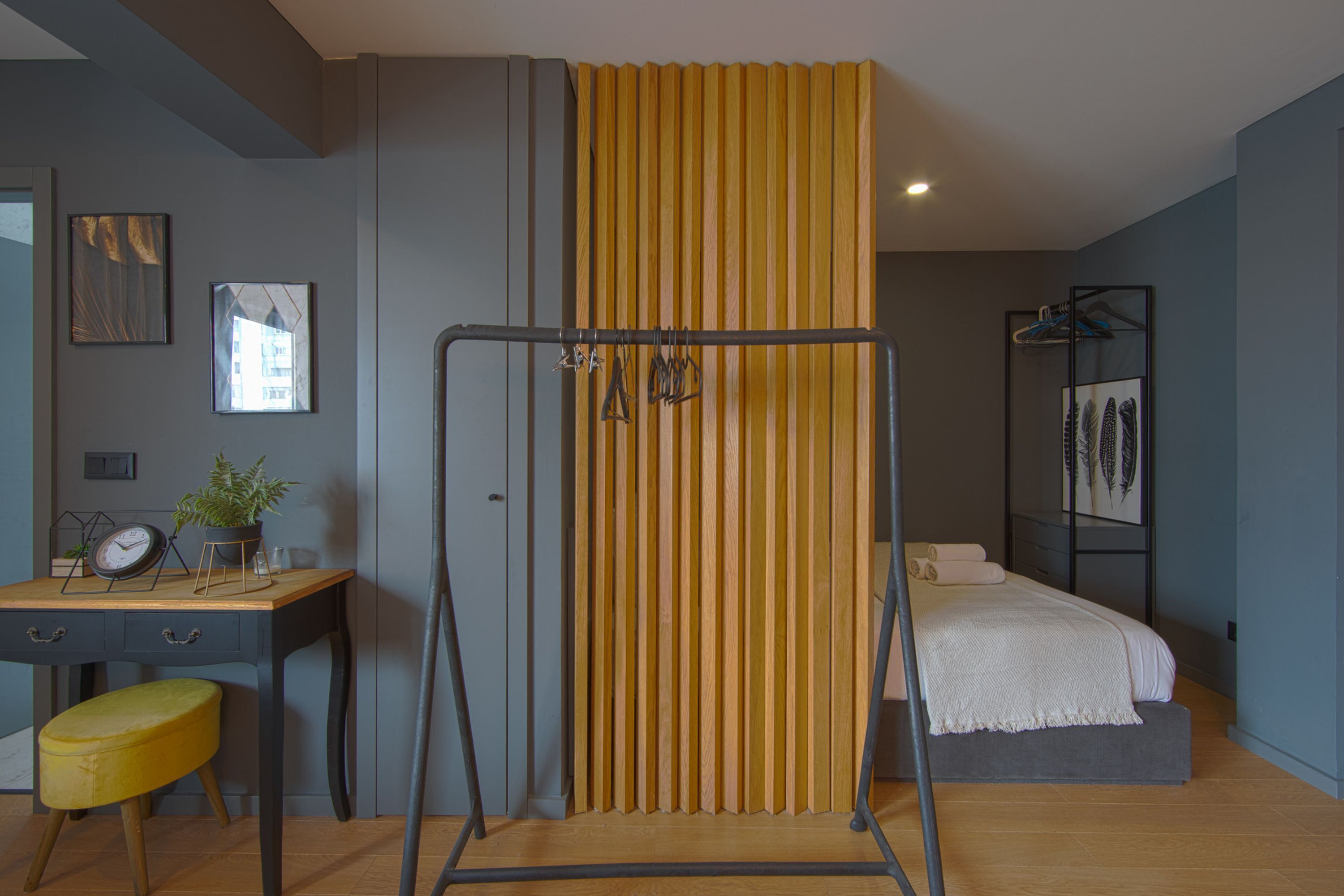 A bedroom with a bed, a desk with a chair, a hanging rack, and a potted plant.
