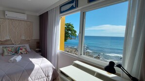 Un dormitorio con cama, mesita de noche, lámpara, ventana con vista al océano y un tapiz de macramé en la pared.