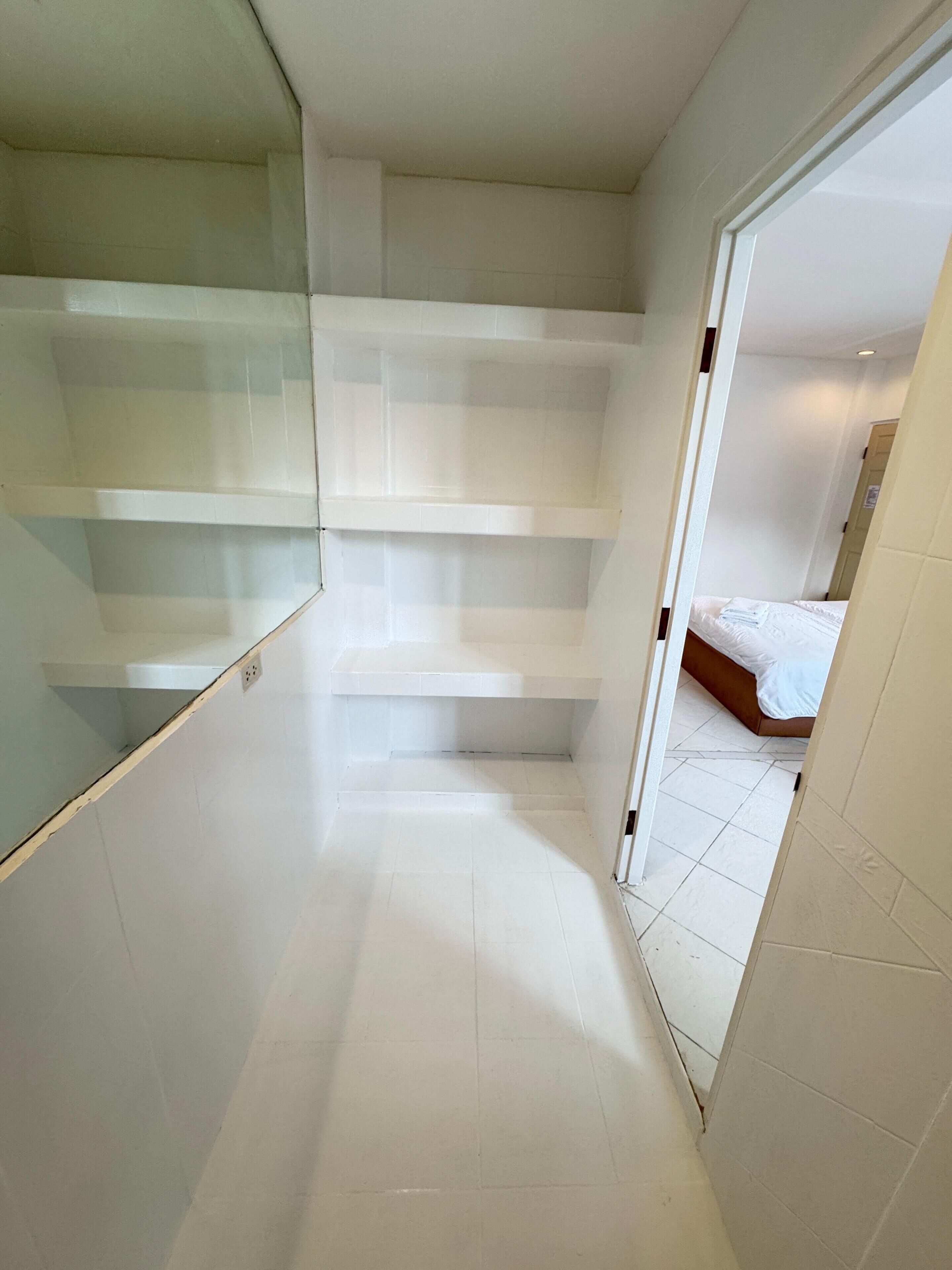 Un couloir étroit aux murs et au sol recouverts de carreaux blancs, qui mène à une chambre à coucher avec un lit et une porte.