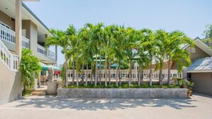 A courtyard with a row of palm trees, a concrete planter, and a building with balconies and a green awning. - Blue Horizon (Hua Hin)
