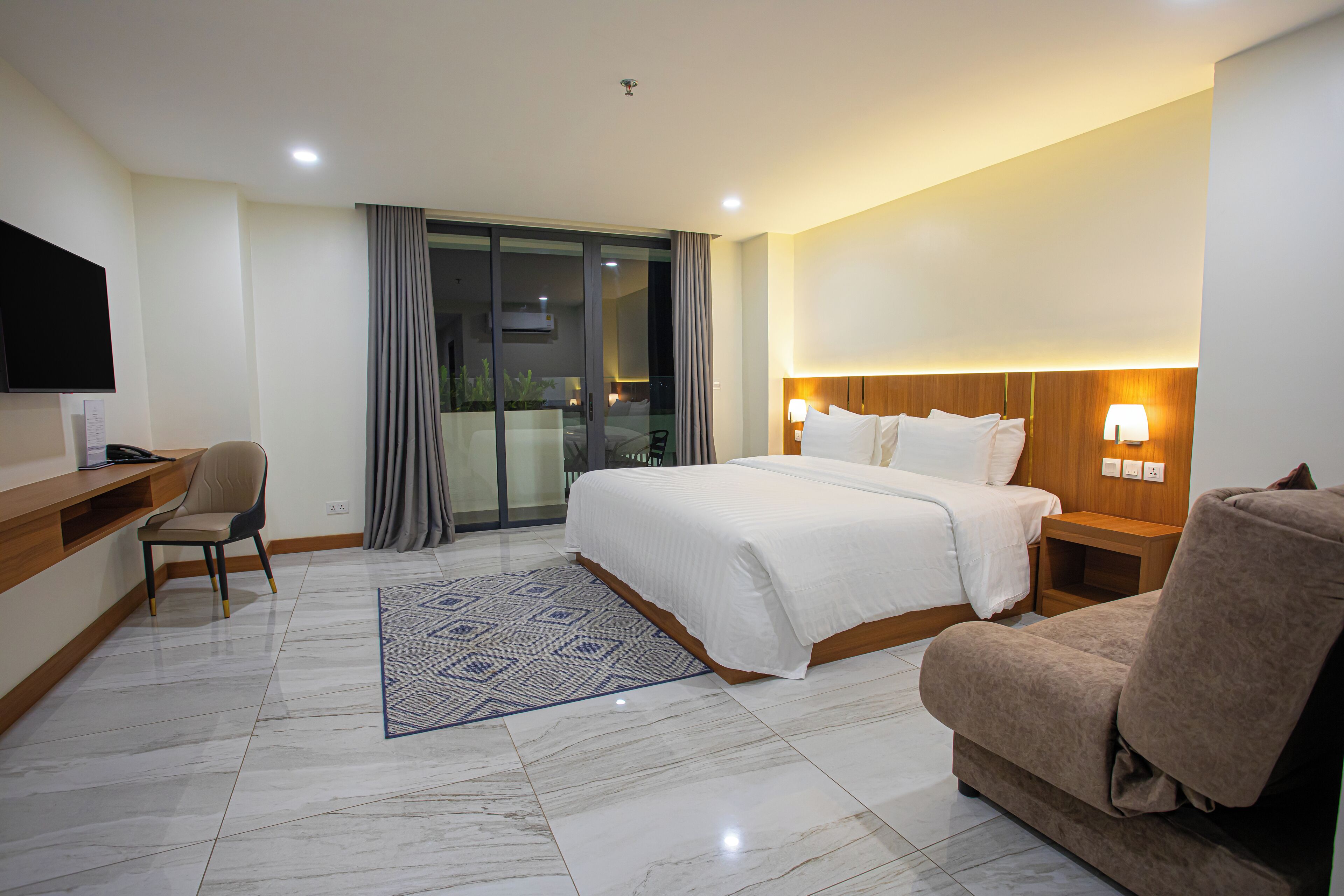 Habitación de hotel con cama, escritorio, silla, televisor y puerta corrediza de vidrio que da a un balcón.
