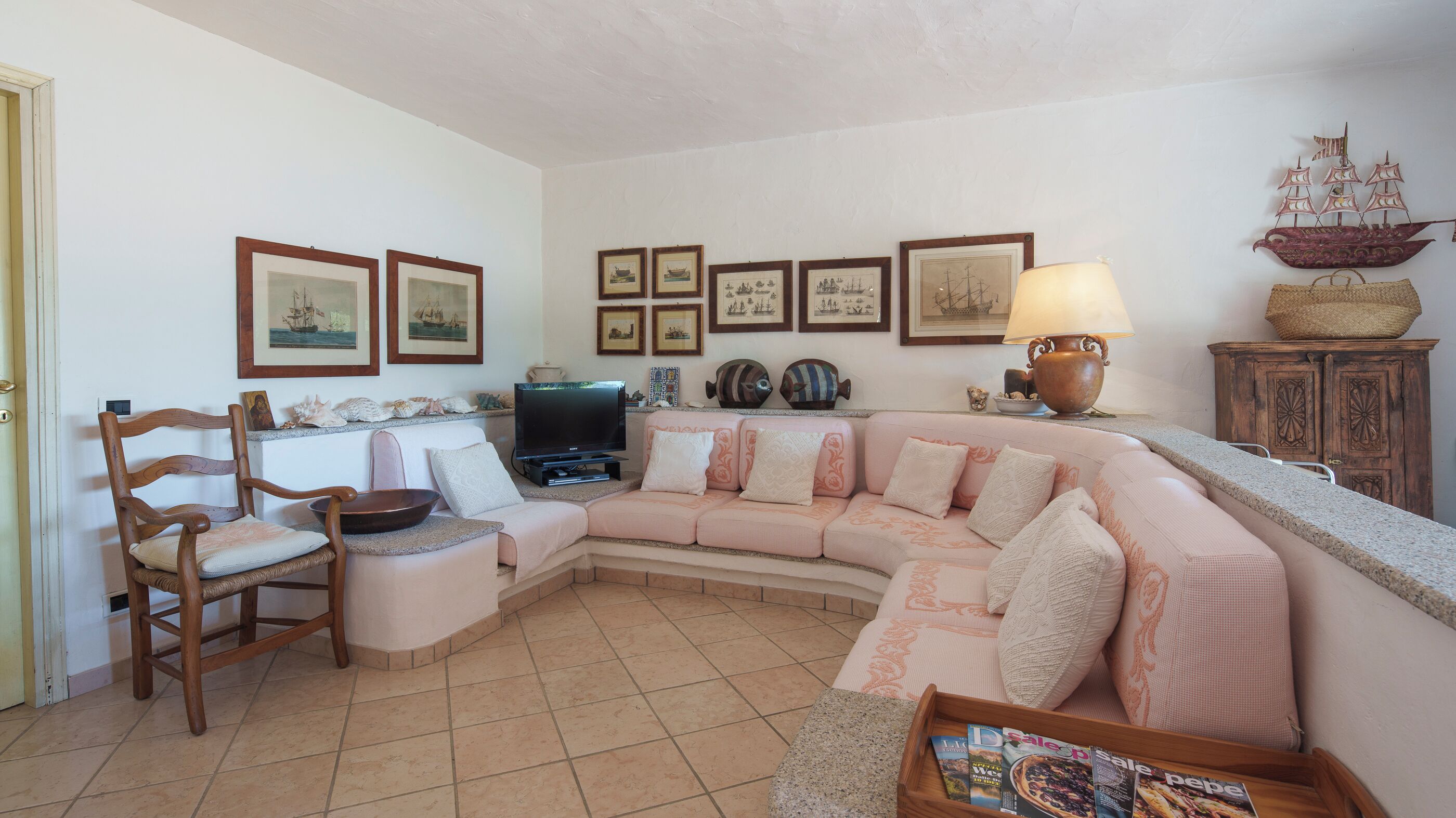 Ein Wohnzimmer mit einer gebogenen Couch, einem Holzstuhl, einem Fernseher, gerahmten Bildern an der Wand und einem dekorativen Schiff auf einem Schrank.
