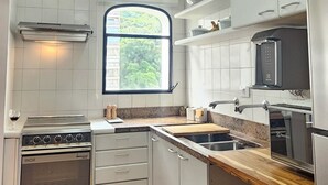 A modern kitchen with white cabinetry, a built-in oven, a microwave, and a window providing natural light. - Pitangueiras Mar Azul 🌊🌴 your cozy beachfront in Guarujá. (Barra Funda Guarujá)