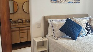 A bedroom with a bed, a nightstand, a wall-mounted air conditioner, a painting, and a bathroom visible through an archway. - Pitangueiras Mar Azul 🌊🌴 your cozy beachfront in Guarujá. (Barra Funda Guarujá)