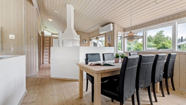 Een moderne eetruimte met een houten tafel en stoelen, een witte scheidingswand en grote ramen met uitzicht op groen.