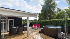 Een houten terras met een jacuzzi, tuinmeubilair en een overdekte zithoek.