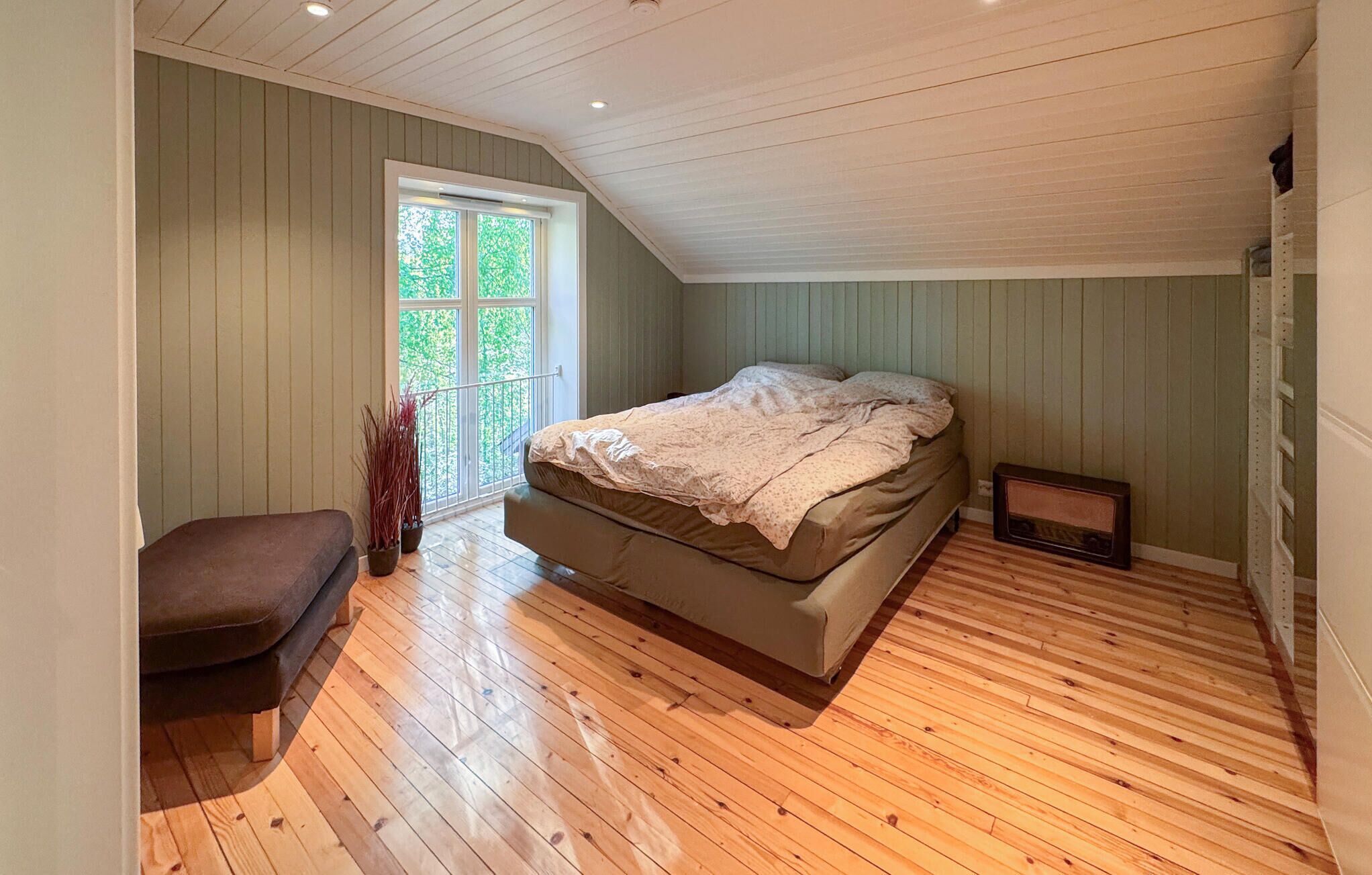 Une chambre avec un plafond en pente, un lit, un banc, une fenêtre et un radiateur.
