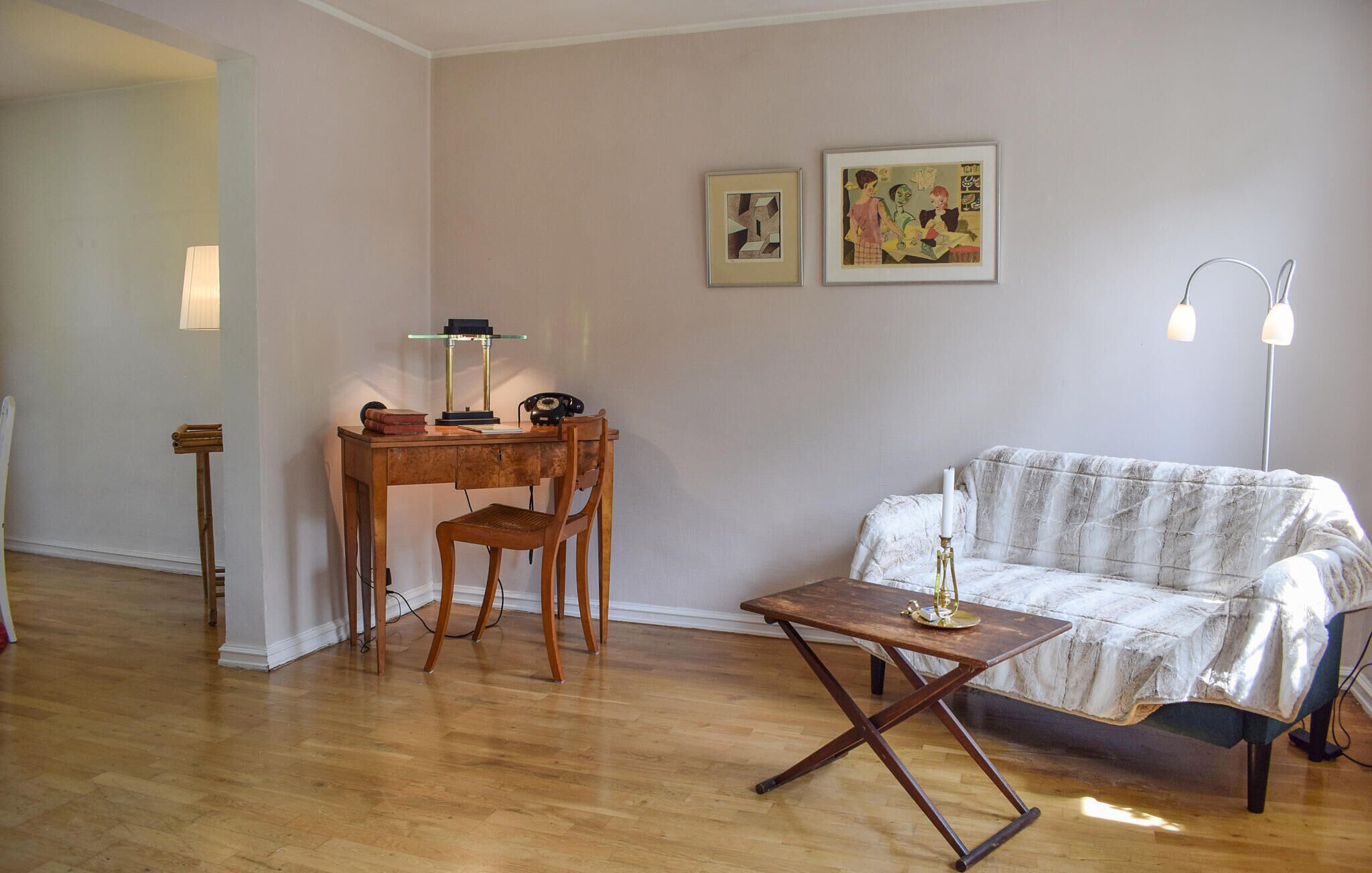 A living room with a wooden desk, a chair, a small table, a sofa, and two framed artworks on the wall.
