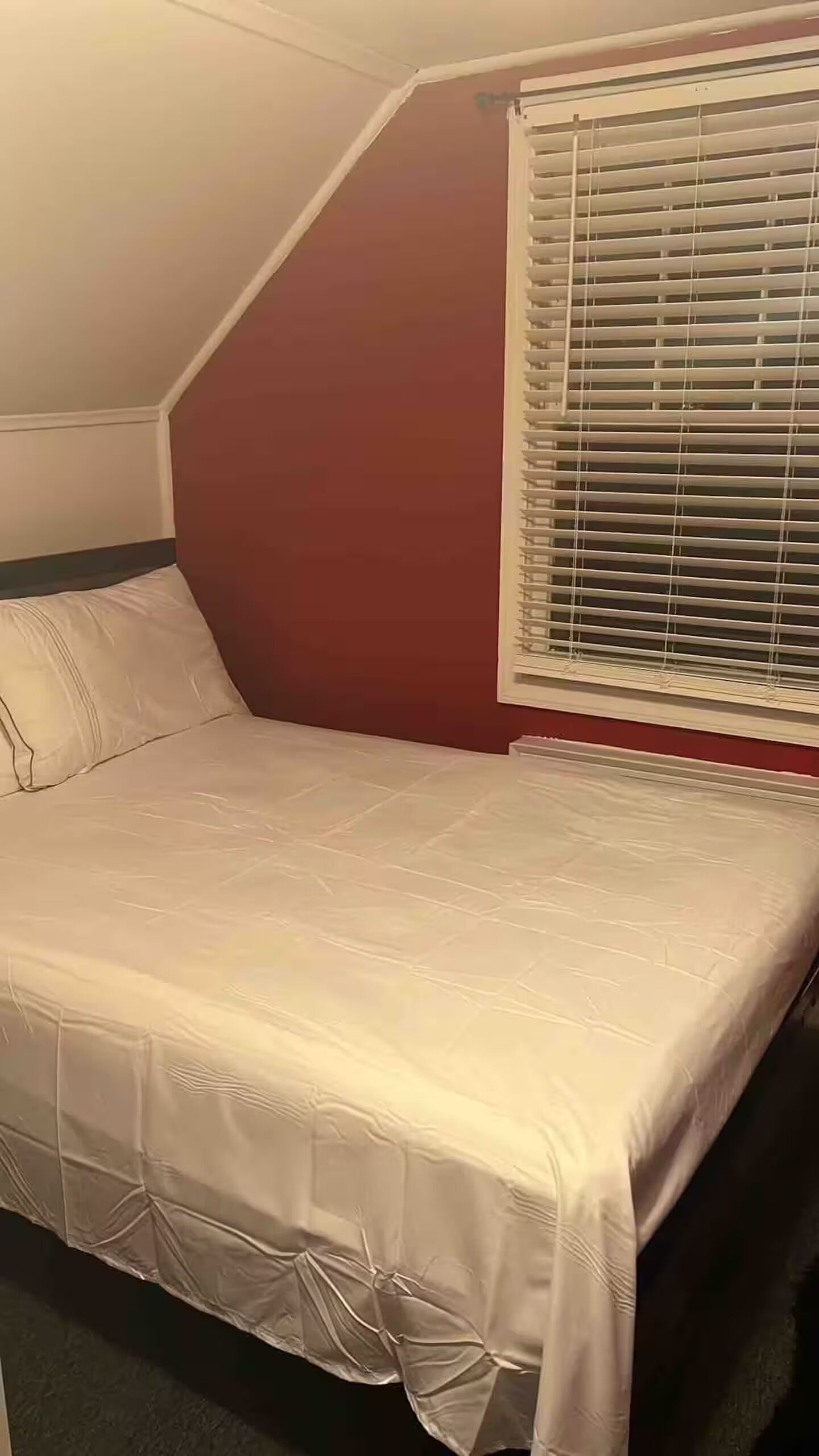 A bedroom with a bed, a window with blinds, and a red wall.