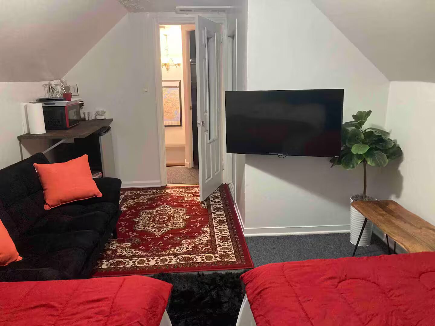 A small, cozy room with a black sofa, red cushions, a red rug, a flat-screen TV mounted on the wall, and a small wooden table.