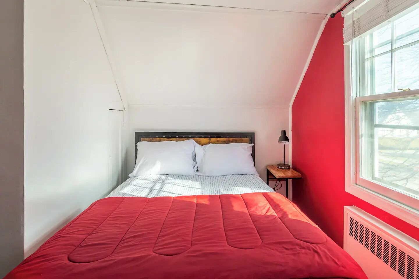 A bedroom with a bed, a nightstand, a window, and a red wall.