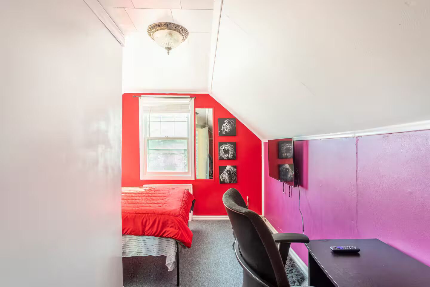 A small, brightly colored room with a red wall, a window, a bed, a chair, and a desk with a remote control.