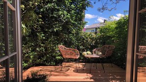 Un patio al aire libre con dos sillas de mimbre y una mesa redonda pequeña, rodeado de abundante follaje y una terraza de madera.