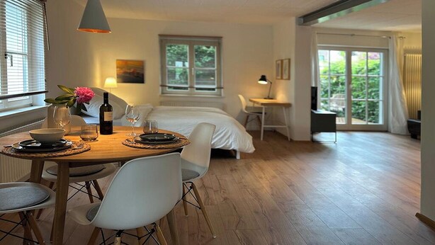 Un dormitorio moderno con cama, mesita de noche y ventana. Hay un comedor con mesa, sillas y un florero con flores.