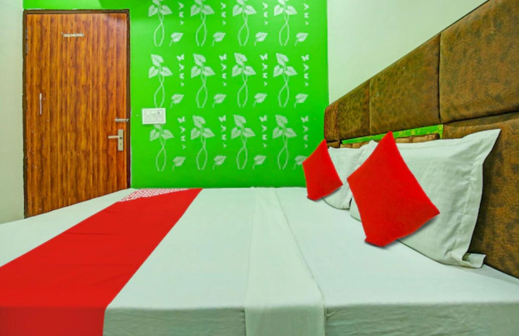 Quarto de hotel com porta de madeira, parede verde com desenhos de plantas em branco, cama com roupa de cama branca e vermelha, e um cobertor vermelho sobre a cama.