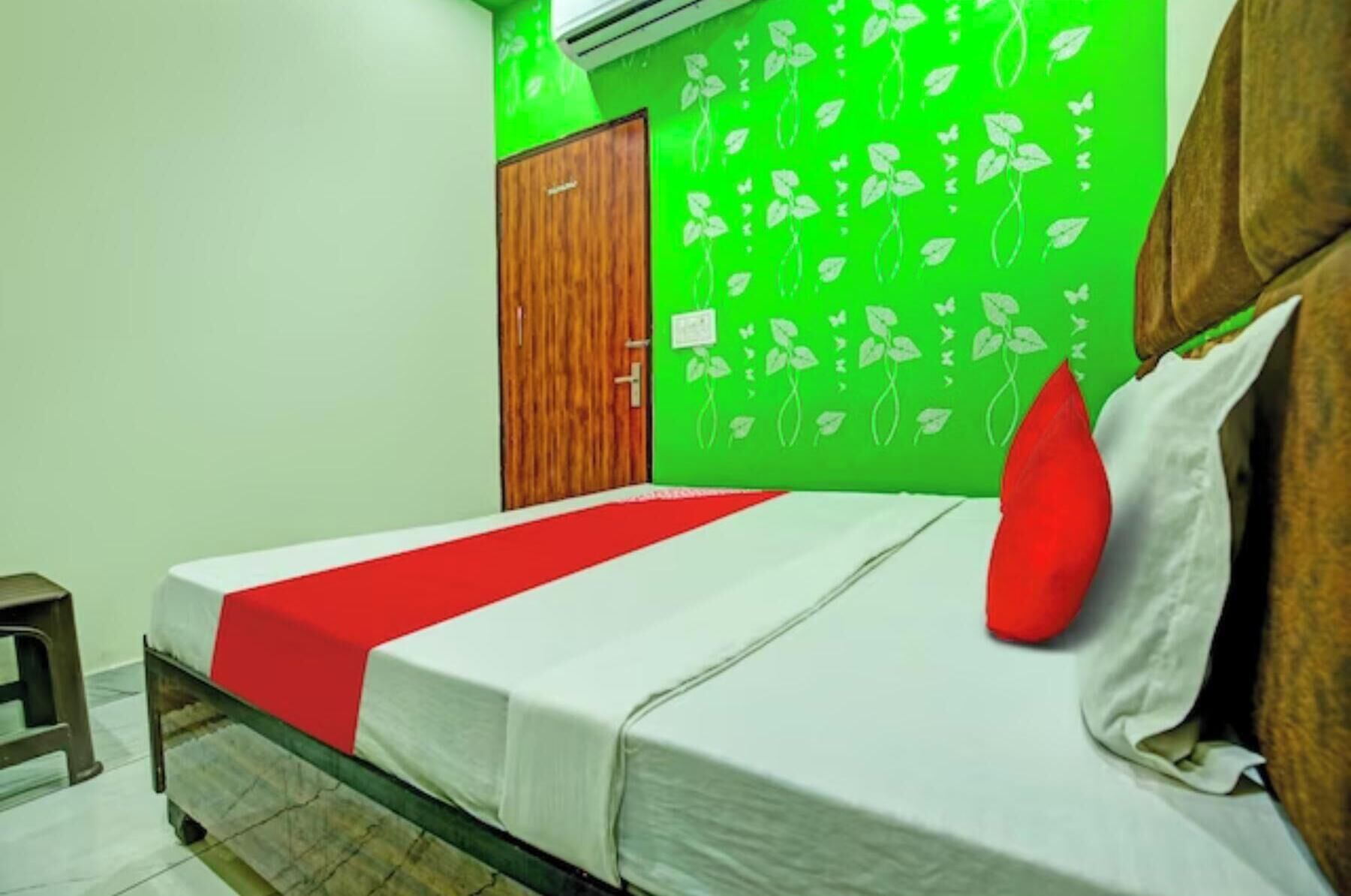 Een slaapkamer met een bed, een rode kussen, een groene muur met bladmotieven, een houten garderobe en een klein krukje.