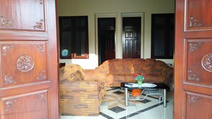 An open wooden door leading to a living room with a sofa, a small table with a vase, and a patterned rug. - Green Surabaya Homey (Surabaya)