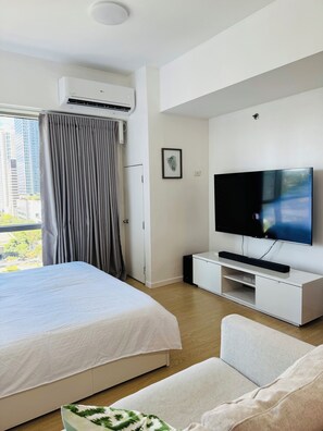 A hotel room with a bed, a TV on a white stand, a grey curtain, and a city view through the window. - Rare King Bed corner unit with City View (Taguig)