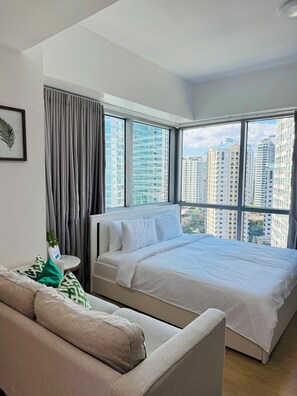 A modern bedroom with a large bed, a sofa, and a view of high-rise buildings. - Rare King Bed corner unit with City View (Taguig)