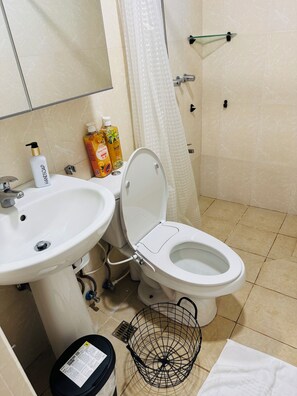 A bathroom with a white sink, toilet, and shower curtain. There are two bottles of hand soap and a bottle of shampoo on the sink. - Rare King Bed corner unit with City View (Taguig)