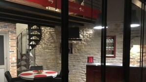 A dining area with a round table and chairs, a brick wall, and a staircase leading to an upper level. - L'aubergiste (Corcelles-les-Arts)