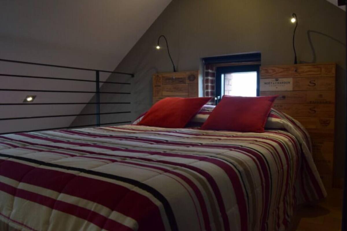 Une chambre avec un plafond en pente, un lit avec une literie à rayures, deux oreillers rouges, une commode en bois et une petite fenêtre donnant sur un beau paysage.