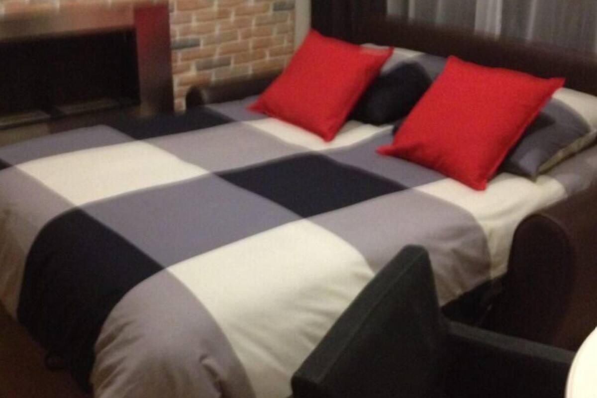 Une chambre d’hôtel avec une couvre-lit à carreaux, deux oreillers rouges, une tête de lit sombre et un mur en briques.