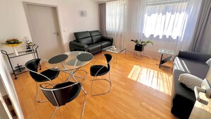 Un salon moderne avec un canapé en cuir noir, une table basse en verre et des chaises noires. On y trouve une petite table d’appoint avec une plante, ainsi qu’une porte donnant sur une autre pièce.