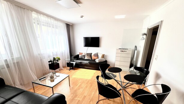 Un salon moderne avec un canapé, une table basse et des chaises noires. Un téléviseur à écran plat est fixé au mur, et il y a une fenêtre avec des rideaux légers.