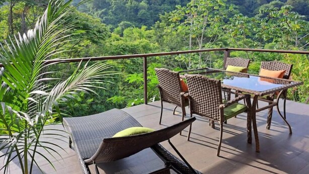 Un balcon agrémenté de meubles en osier, d’une table en verre et offrant une vue sur une forêt luxuriante.