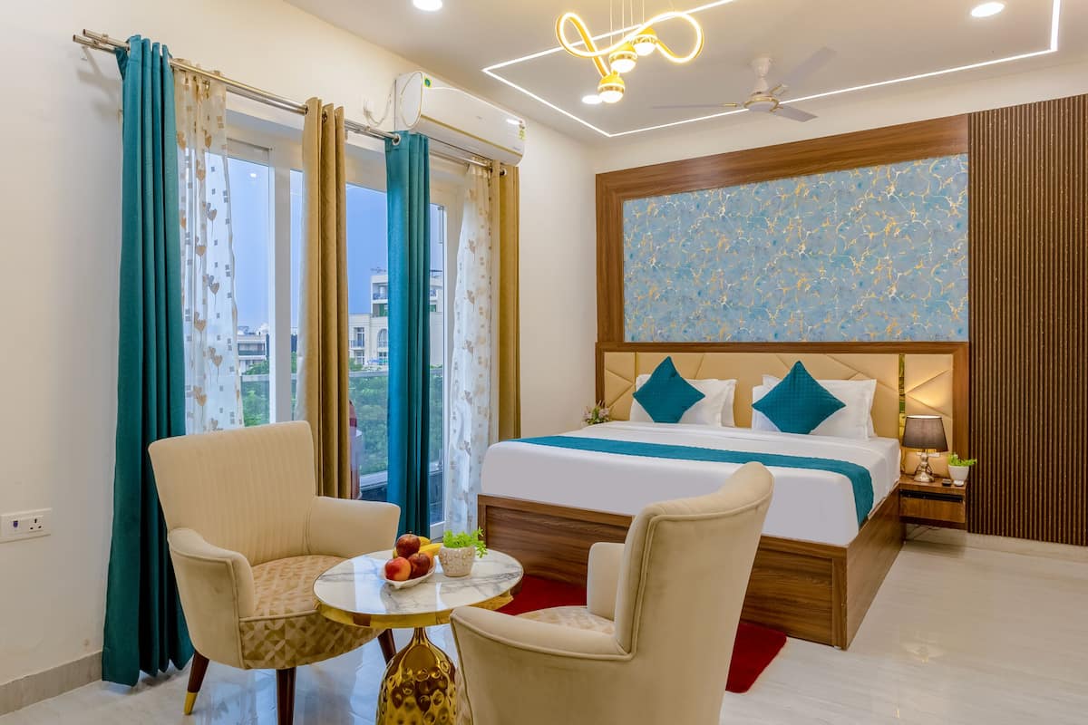 Une chambre d’hôtel moderne avec un grand lit, deux fauteuils, une petite table avec des fruits et une plante, et une vue sur la ville grâce à de grandes fenêtres.