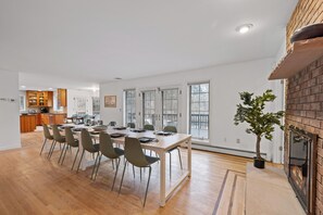 A spacious dining area with a long table and chairs, a brick fireplace, and large windows.