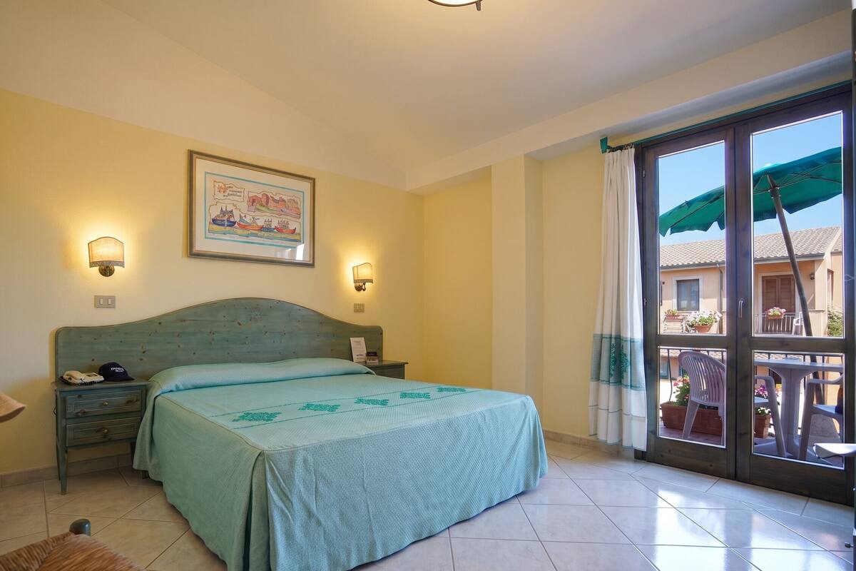 A bedroom with a bed, two wall-mounted lamps, a framed map, and a balcony with a view of the outside.