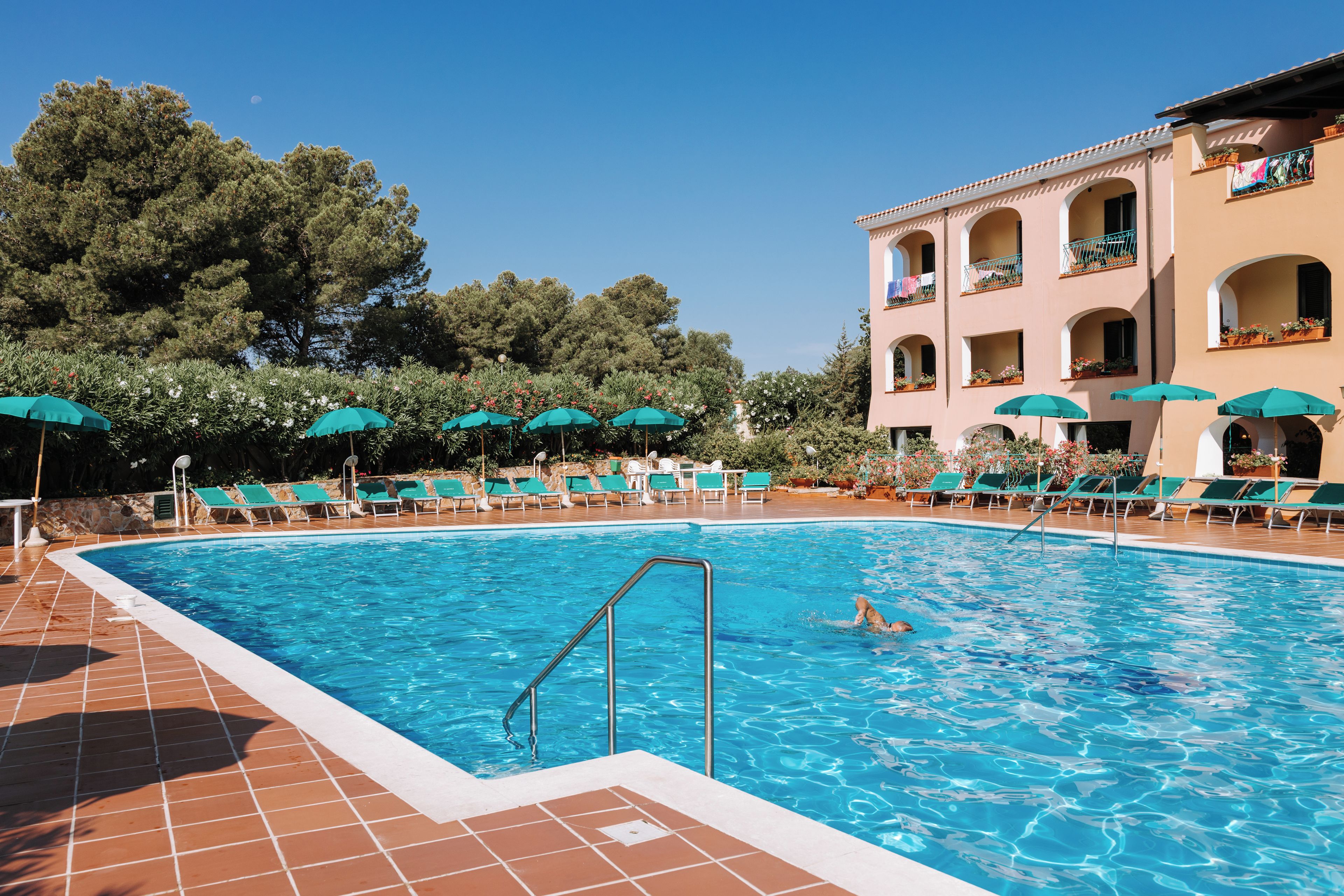 A swimming pool with a tiled deck, surrounded by lounge chairs and umbrellas, and a building with balconies in the background.