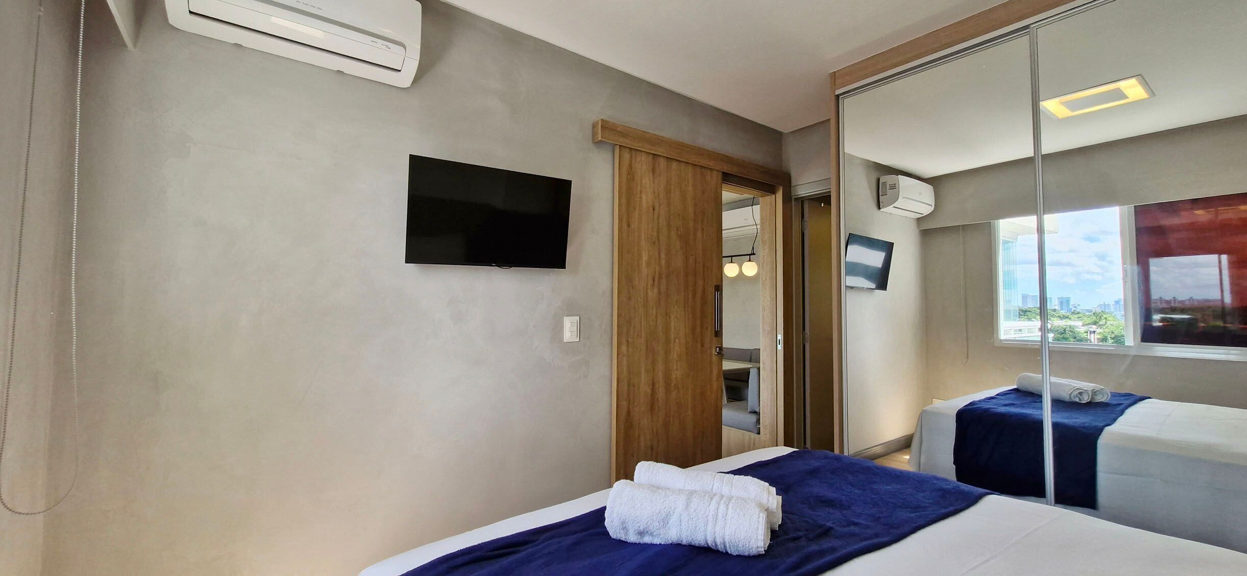A hotel room with a bed, a TV, a mirror, and a window with a city view.