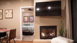 A living room with a fireplace, a flat-screen TV mounted above it, a dining area with a table and chairs, and a bed in the background. - Edelweiss MTN Haus, Summer Base Camp Steps from PCMR, Pool, Hot Tub. Local Hosts (Park City)