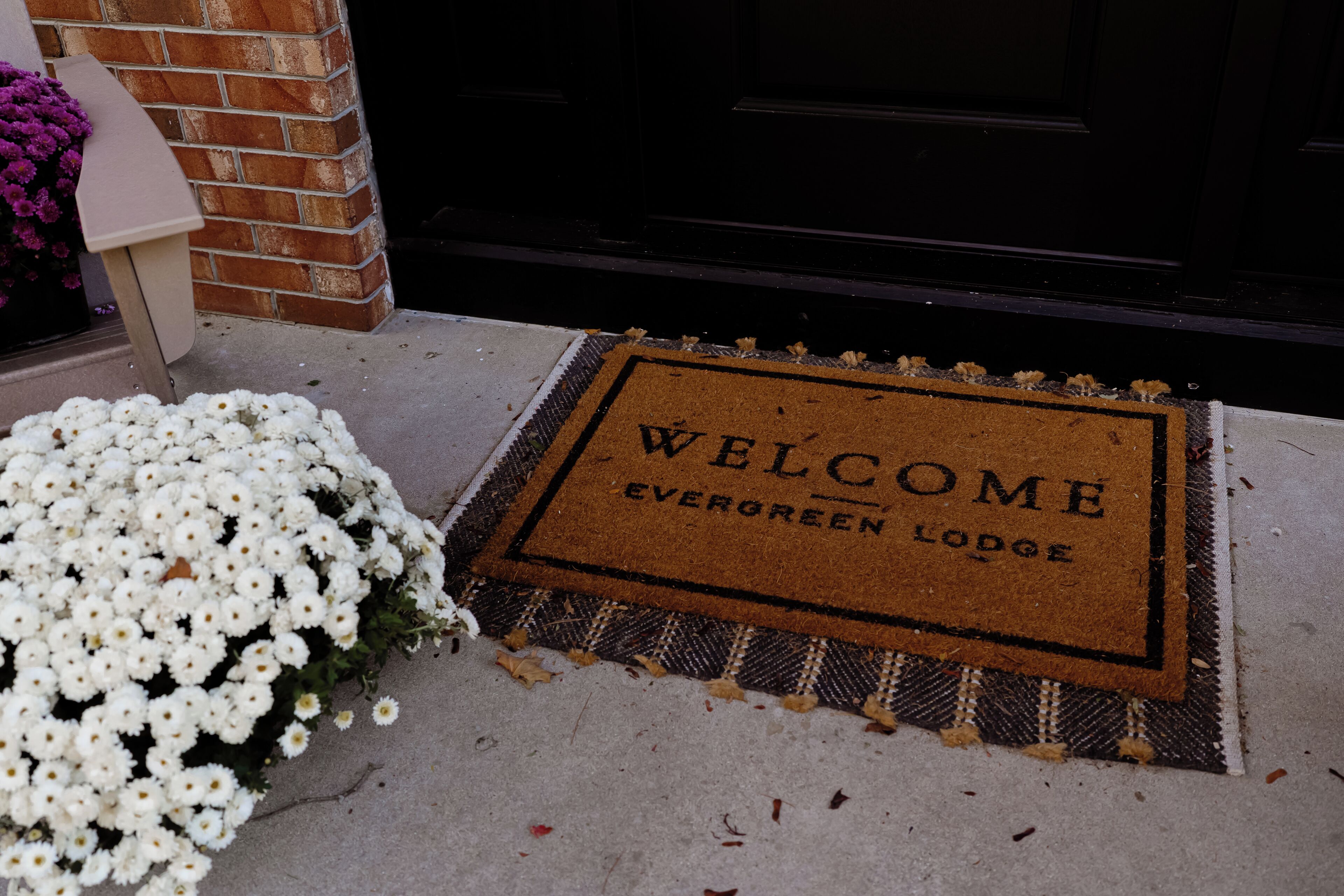 A welcome mat with 'WELCOME EVERGREEN LODGE' text, a brick wall, and a white flower arrangement.