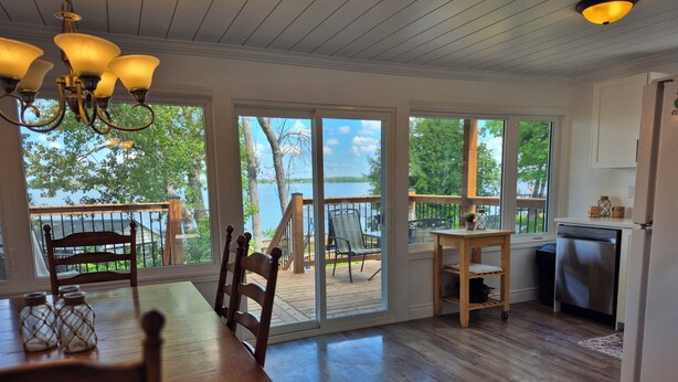 Eine Holzterrasse mit Esstisch und Stühlen, ein Küchenbereich mit Geschirrspüler und große Fenster mit Blick auf einen See und Bäume.
