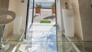 A room with a stone archway leading to a balcony, a glass table with decorative items, and a modern bathroom with a sink and mirror. - SUITE ORTIGIA SEA VIEW (Siracusa)