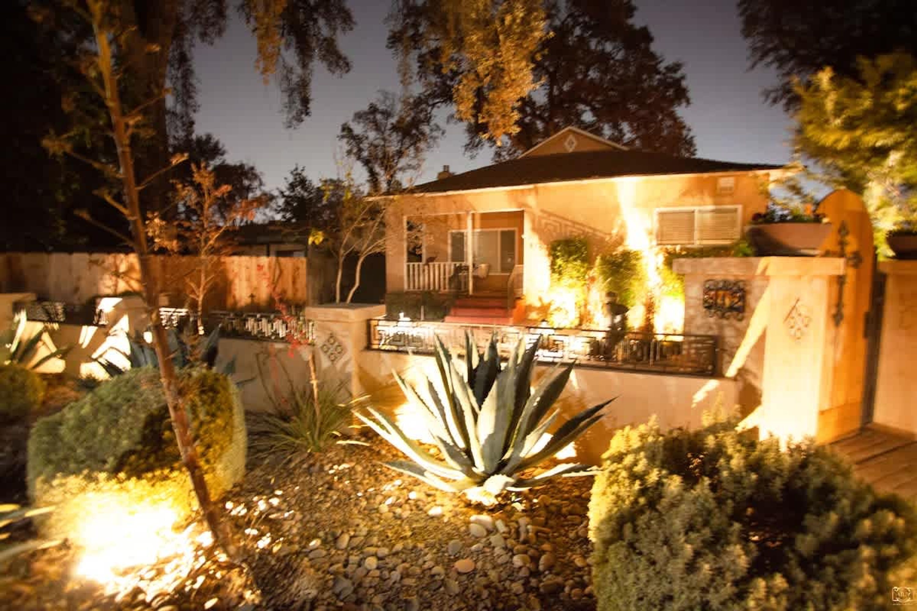 Una casa con porche cubierto, un muro de piedra y un jardín bien iluminado con plantas de agave.