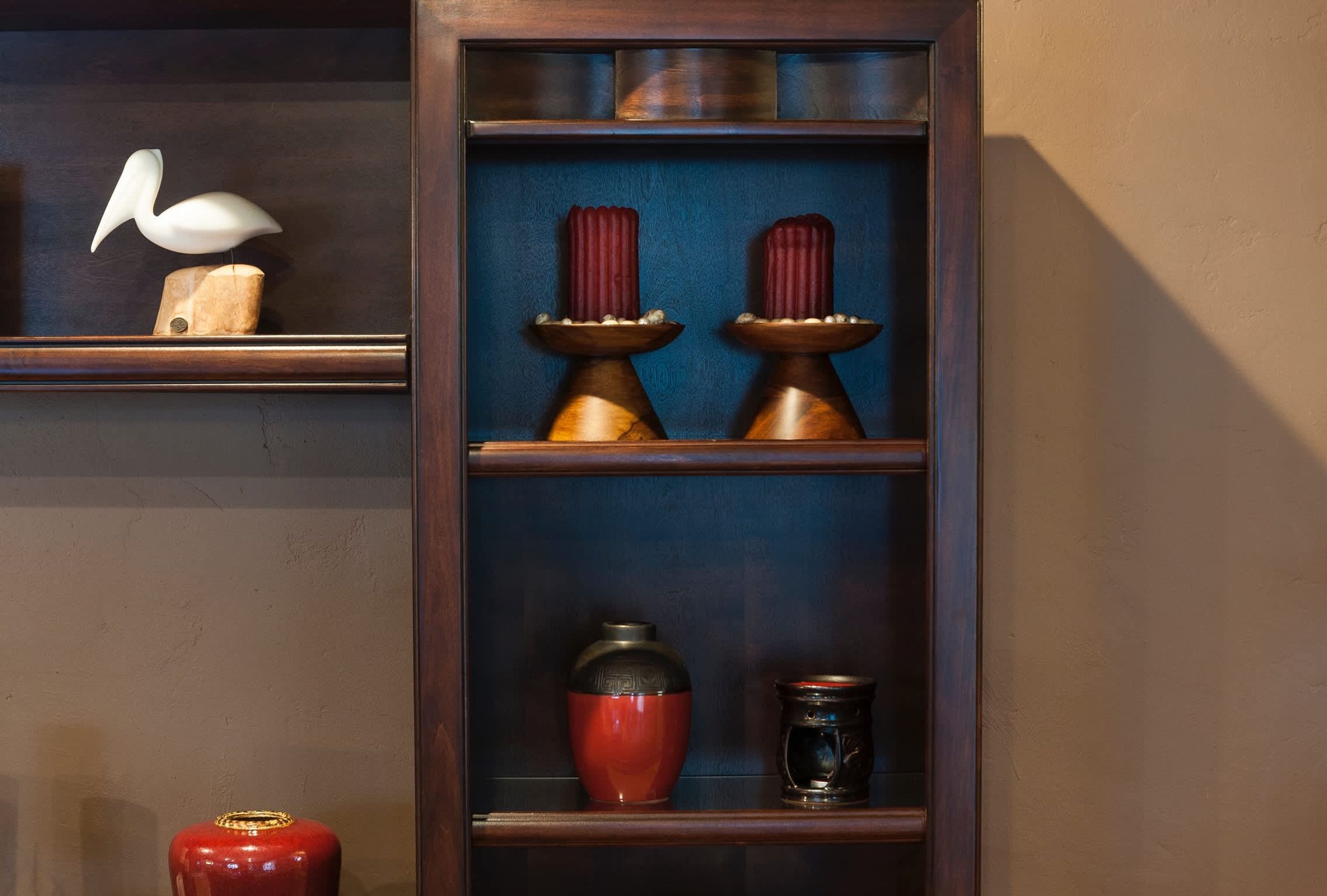 Una estantería de madera con objetos decorativos, incluyendo una escultura de pájaro blanco, dos candelabros, un jarrón rojo y un pequeño recipiente negro.