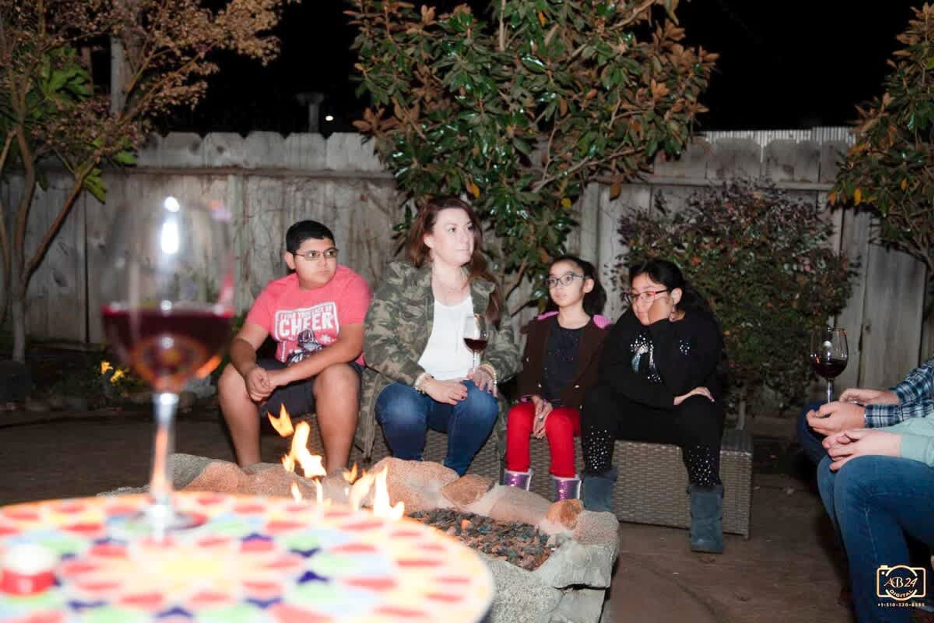 Un groupe de personnes est assis autour d’une table, devant un gâteau et des verres de vin, à l’extérieur, la nuit.